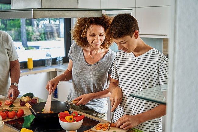 Familie kocht gemeinsam. Während die Mutter rührt, schneidet der Sohn das Gemüse klein.
